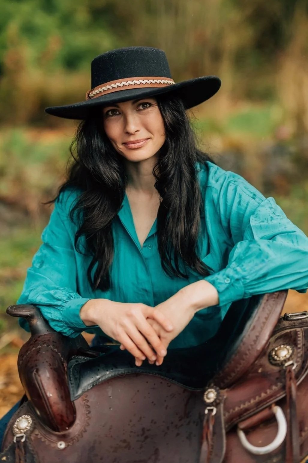 RM Peacock in a black flat-brim hat and teal blouse, one hand resting on a worn leather saddle.